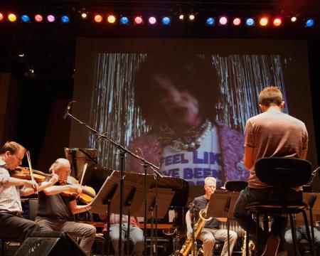 A close up shot of London Sinfonietta musicians and a large screen in the back showing performance artist David Hoyle